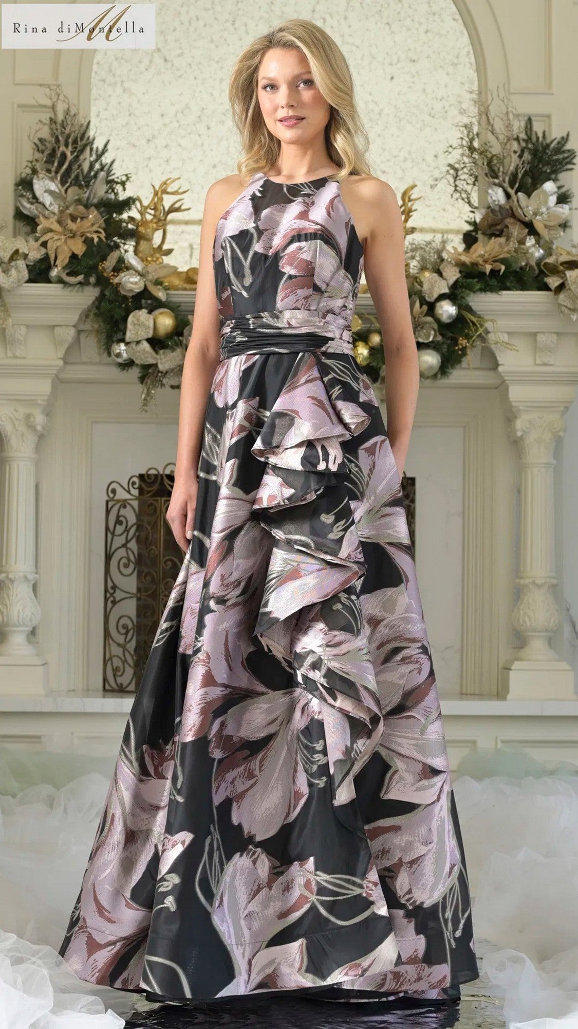 A woman stands indoors in the Rina Di Montella RD3361—a sleeveless, pink multi floral A-line gown with ruffle details—complementing a white fireplace adorned with flowers and greenery.